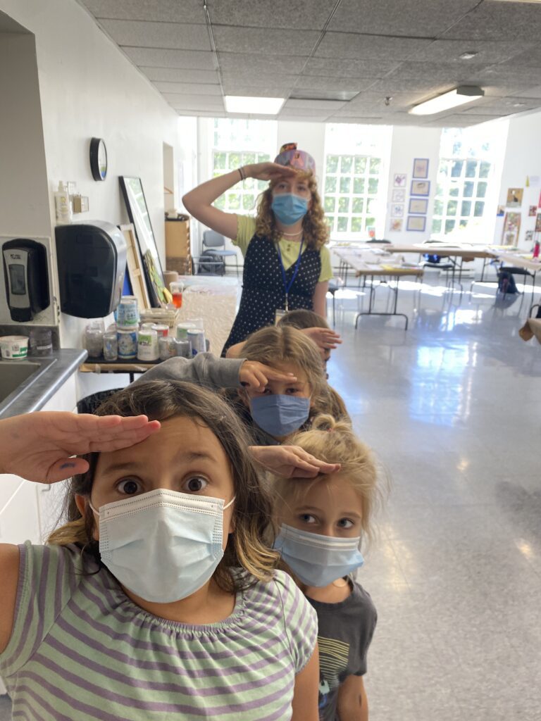 Photograph of four young kids in a straight line with their teacher behind, saluting to the photographer.