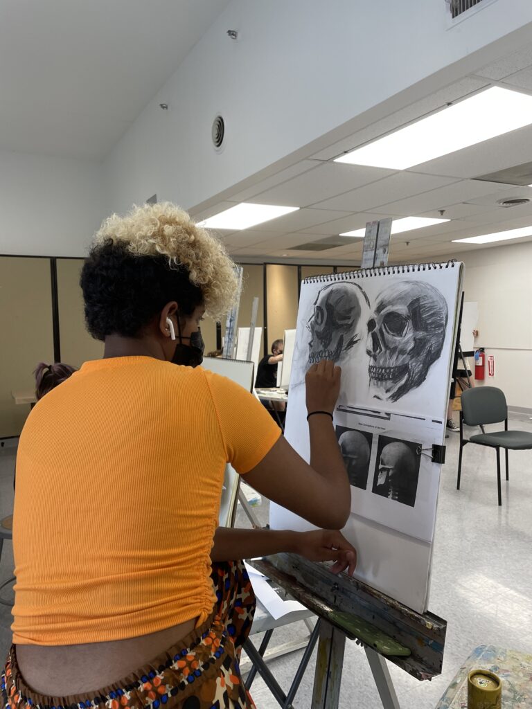 Photograph of a teen in a bright orange shirt seated at an easel with a sketch pad drawing two angles of a human skull.