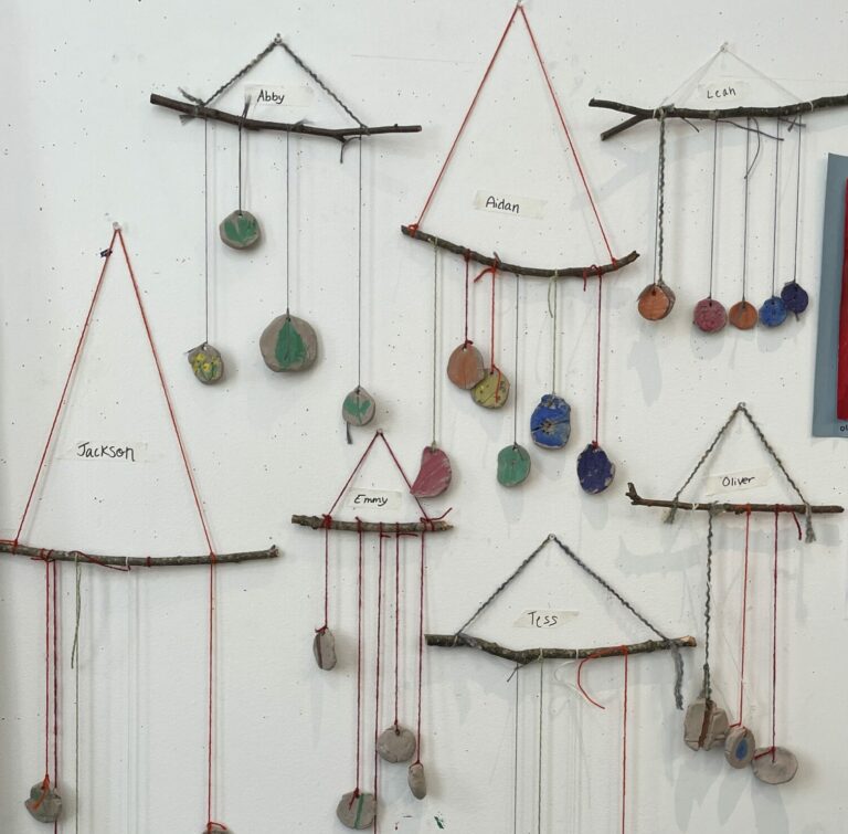 Display of seven hanging sticks with strings attaching various clay shaped objects to each one.