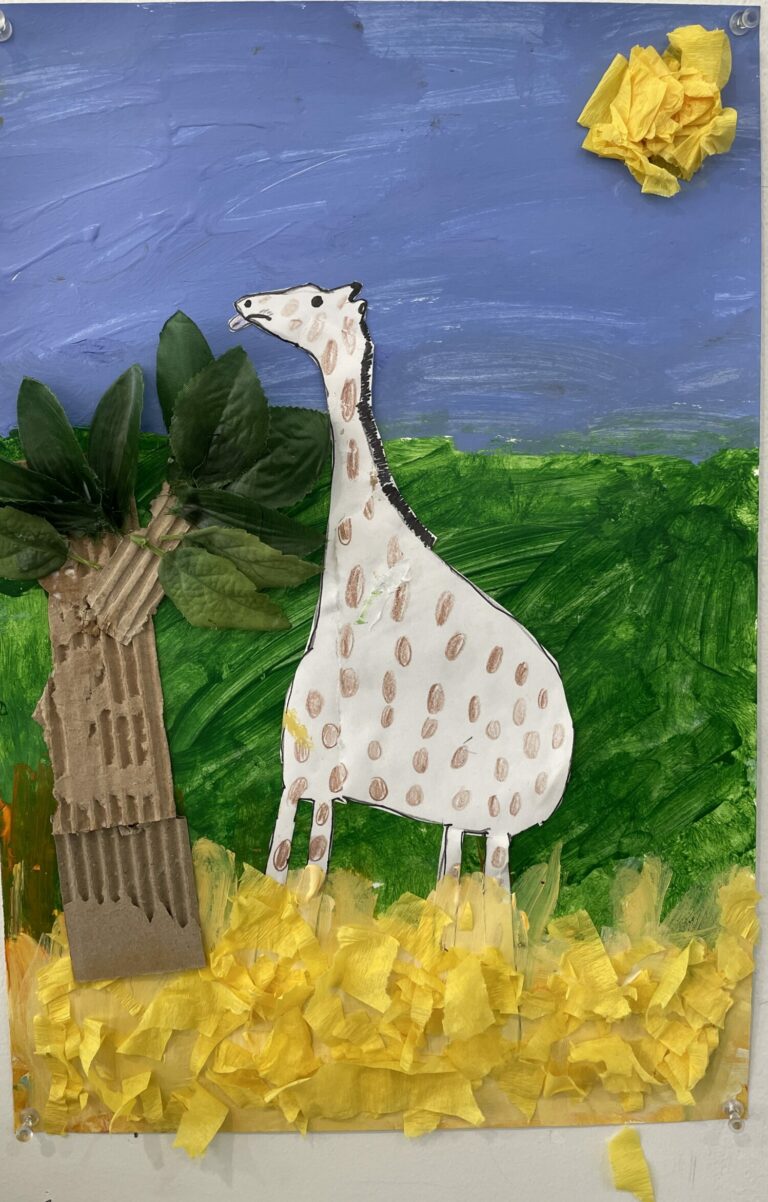 Photograph of a landscape with painting blue sky and green grass. There is a collage tree, yellow paper ground and sun, and a paper giraffe eating from the tree.