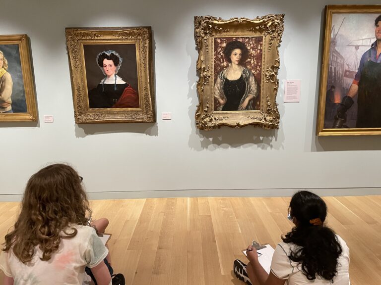 Photograph of two teens seated on the ground in a museum gallery. They are drawing on clip boards from the painting portraits hanging in front of them.
