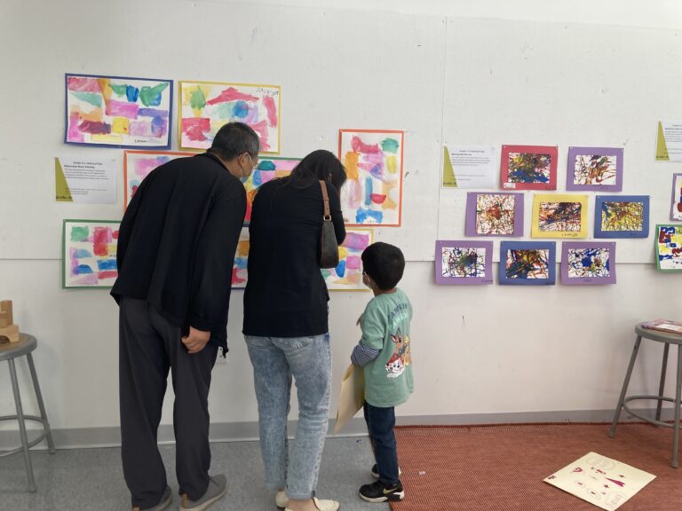 Parents and a child viewing artwork hanging on a white cork wall.