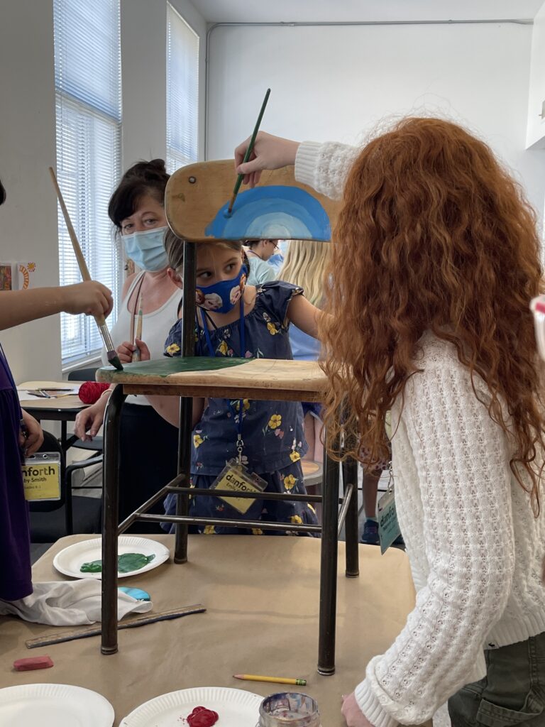 Photograph of a group of young students painting a chair with blues and greens.