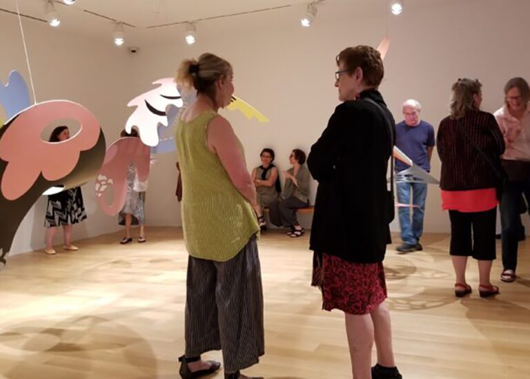 Photograph of a group of people gathered around an installation hanging from the ceiling. In the foreground are two women with backs towards the camera talking.
