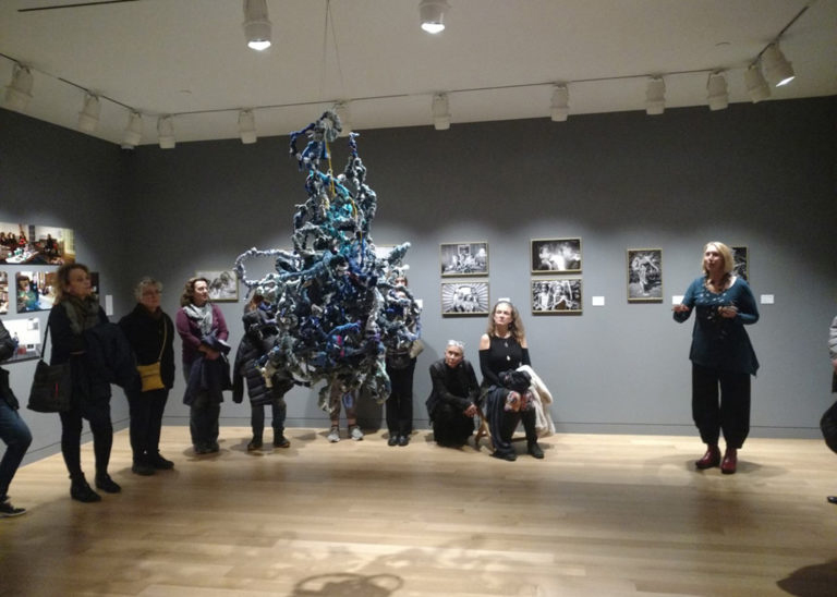 Photograph of a lecture in a gallery space. The gallery has dark grey walls and tan wood floor. In the center hanging from the ceiling is a ball of blue squiggled fabric. On the walls are various photographs. There are people standing in an arch around the ball.