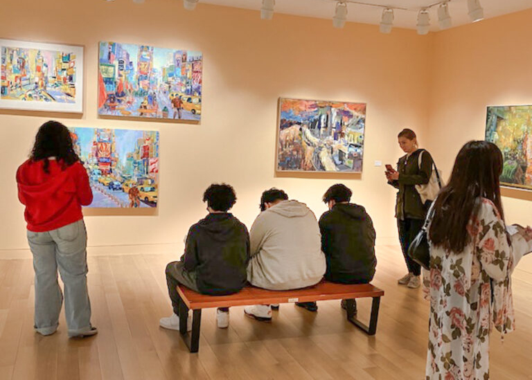 Group of teenagers in a museum gallery with peach walls and busy paintings. Three students stand around a group of three students seated on a bench.