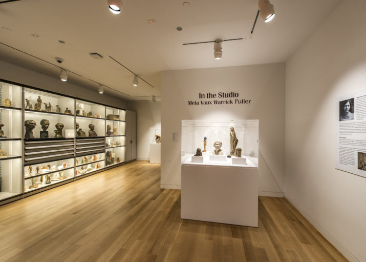 Photograph of a gallery room with white walls and light wood floor, There is a large pedestal with five sculptures. On the far left is a whole wall of lit plexi covered cases filled with sculptures.