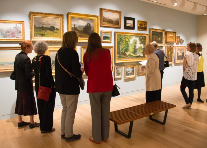 Photograph of a group of visitors in a museum looking at a vast number of paintings on a light blue gallery wall.