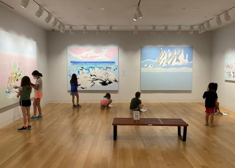 Photograph of a group of children in a museum gallery with four large surreal paintings on the walls. The children are using the works as inspiration.
