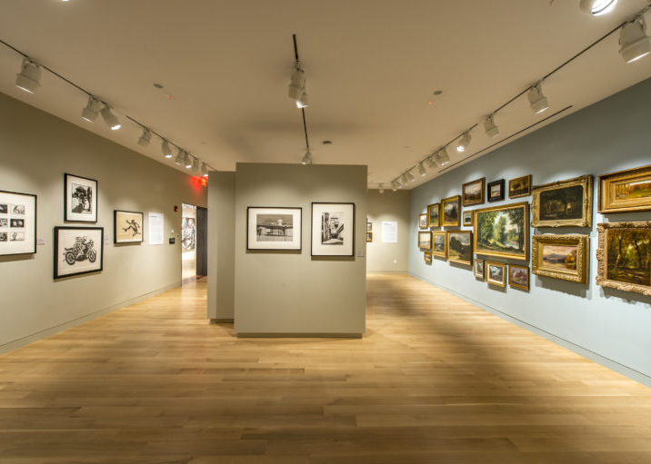 Photograph of a gallery room with light blue and tan walls and light wood floor. There are various pictures on all walls, including many 19th century works in gold frames on the right side hung close together at different levels.