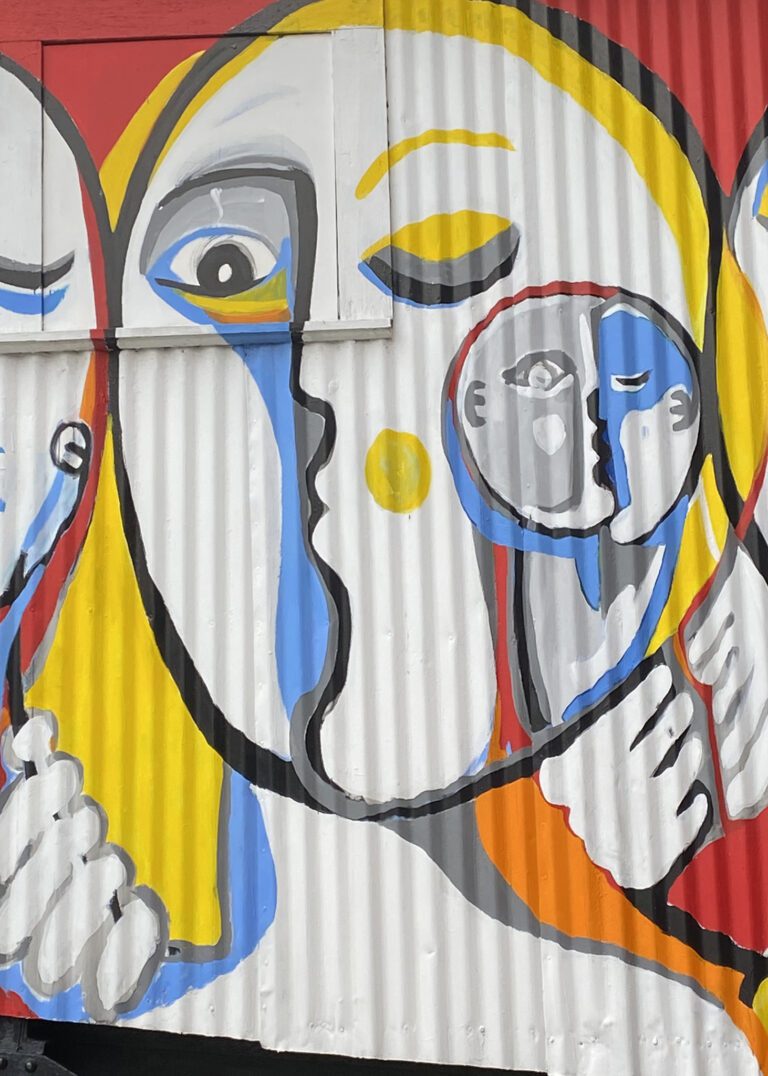 Close up of a mural on a metal corrugated surface. The image is of an oval abstract face with another smaller face on its right cheek. The faces are outlined in black with additional colors of yellow and blue.