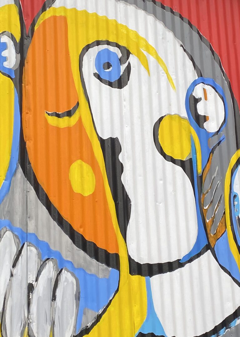 Close up of a mural on a metal corrugated surface. The image is of an oval abstract face. The face is outlined in black with additional colors of yellow and blue, with its left side all in orangy-red.