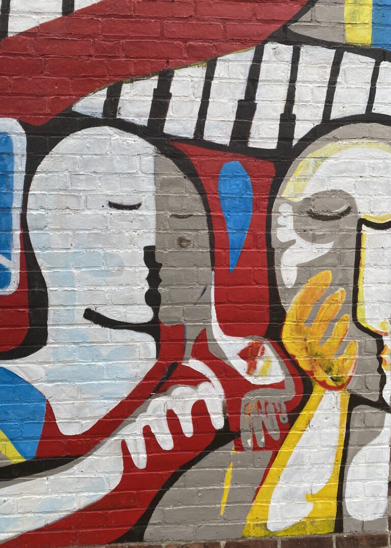 Photograph of a brick wall with a mural. The mural is abstract figures in shades of white with highlights of blue, red, and yellow.