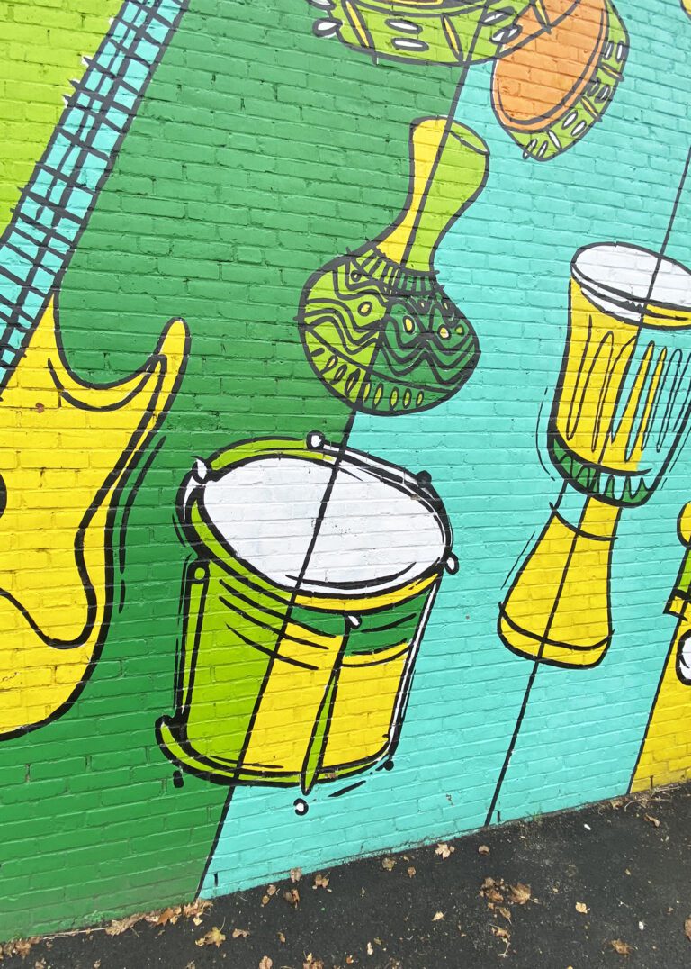 Photograph of a brick wall with a brightly colored mural of diagonal lines in different shades of green and yellow. Also going diagonally are various instruments painted to compliment the background colors, including drums, tambourines, and guitar.