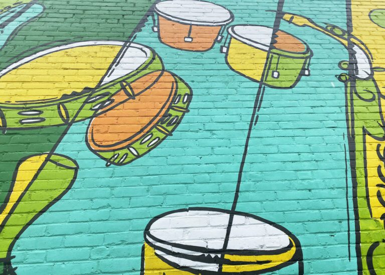 Photograph of a brick wall with a brightly colored mural of diagonal lines in different shades of greens and yellow. Also going diagonally are various instruments painted to compliment the background colors, including drums, tambourines, and saxophone