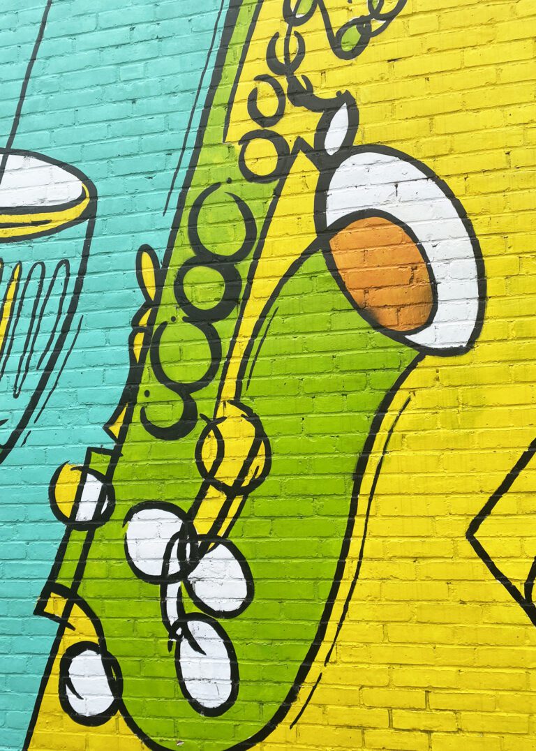 Photograph of a brick wall with a brightly colored mural of diagonal lines in different shades of greens and yellows. Also going diagonally are various instruments painted to compliment the background colors including drums and saxophone.