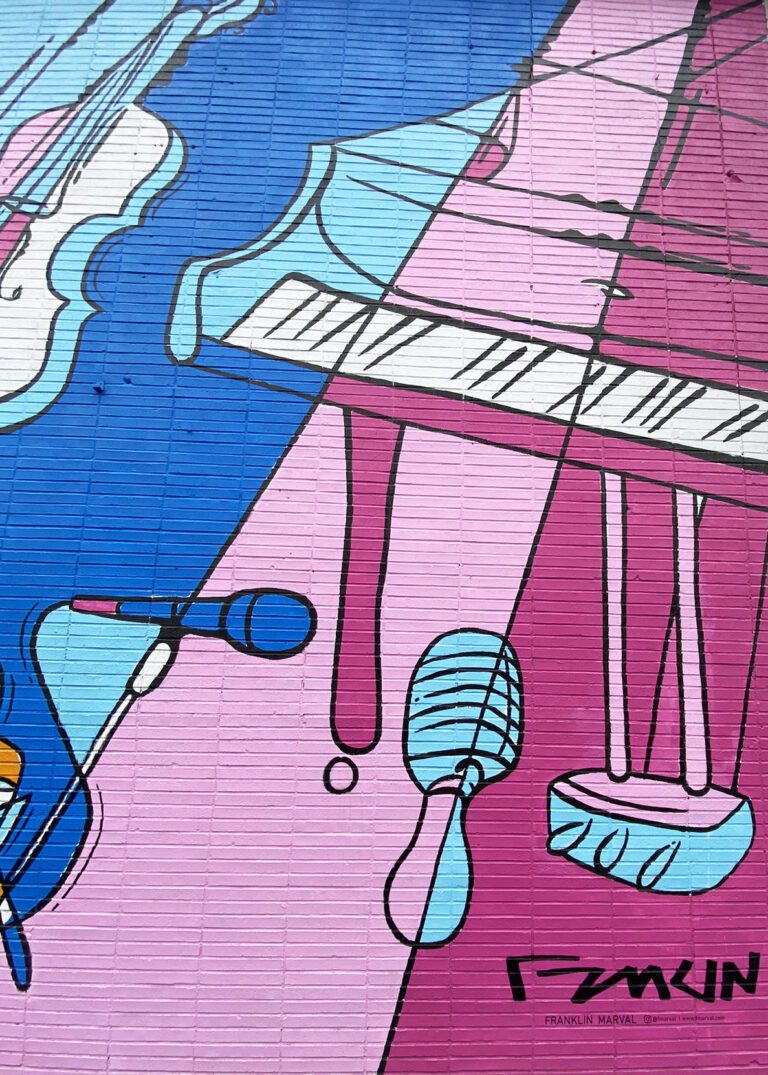 Photograph of a brick wall with a brightly colored mural of diagonal lines in blues and pinks. Also going diagonally are various instruments painted to compliment the background colors, including a piano, microphone, and violin.