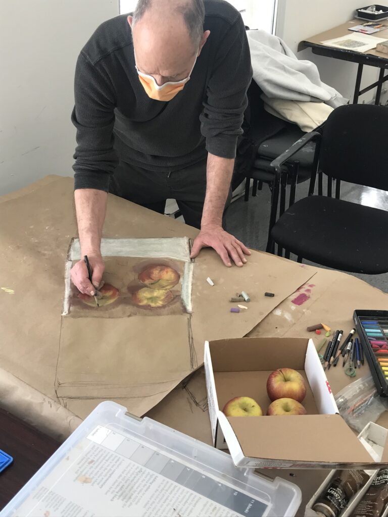 Photograph of a man standing at a table, drawing on brown paper a box of apples from a still life.