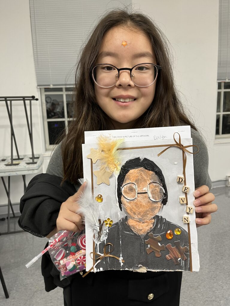 Photograph of a girl with long straight hair and glasses. She is holding a drawing and collage self portrait .