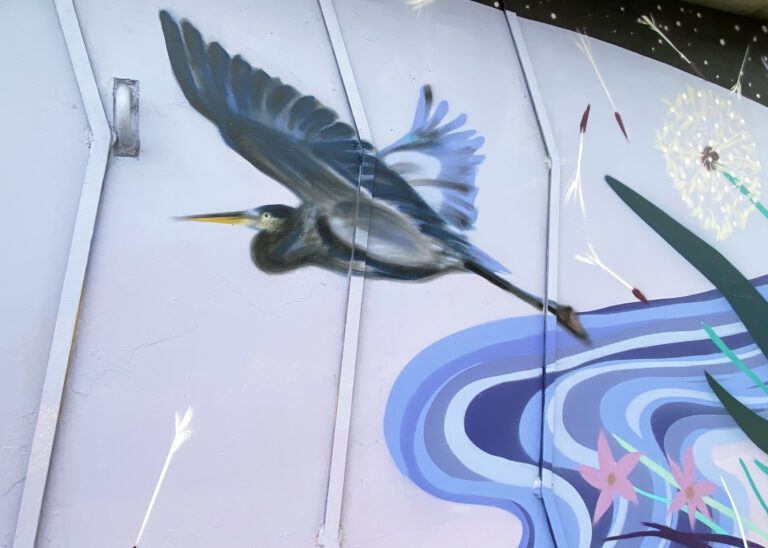 Close up of a mural with a pinky-blue background and a swirling pattern with shades of blue on the bottom right. In the center is a heron flying. To the upper right is a dandelion with seeds blowing away.