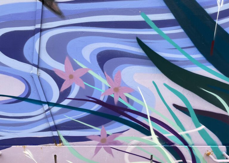 Close up of a mural with a swirling shades of blue pattern behind blades of grass in various shades of green with three pink flowers.