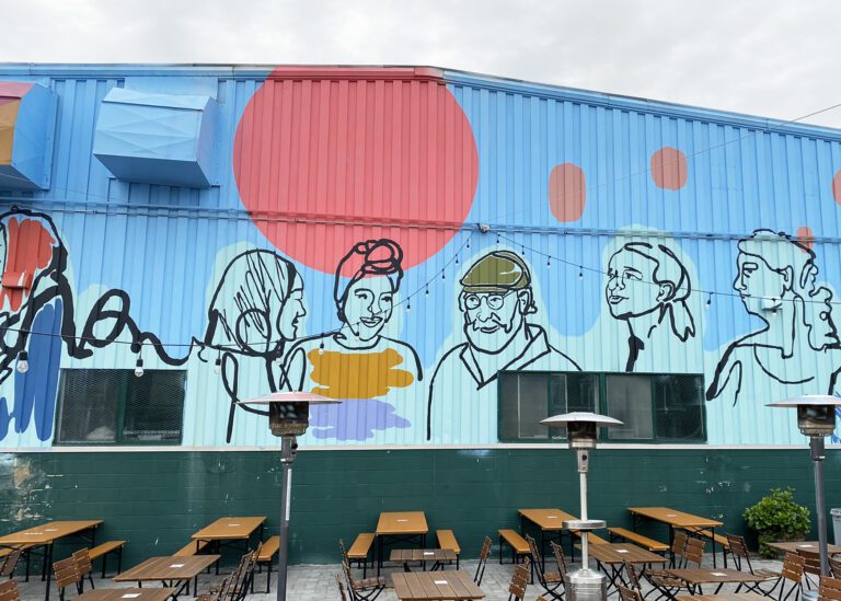 Photograph of a metal corrugated building in a bright blue. There is a mural with black outlines of people painted in a lighter blue. Above them is a bright pink circle.