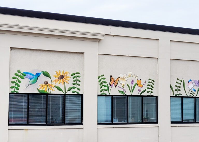 Photograph of a tan building with murals painted above the squat windows of green stems, various flowers, butterflies, and songbirds.