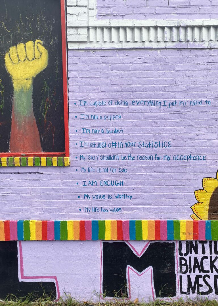 Close up of a mural with a purple background, a yellow, green and red fist in the upper left, and a rainbow pattern across the bottom. There is writing in both blue and black across the surface.