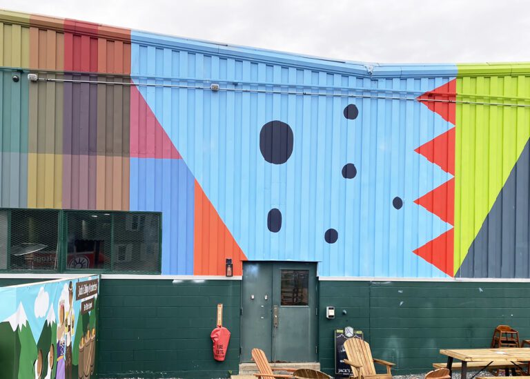 Photograph of a metal corrugated building in a bright blue. There is a mural with bright shapes and black dots.