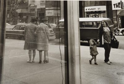 Black and white photograph at a glass building corner. There is a wide sidewalk with a woman holding a child's hand looking back at two older women arm in arm smiling at the young girl.