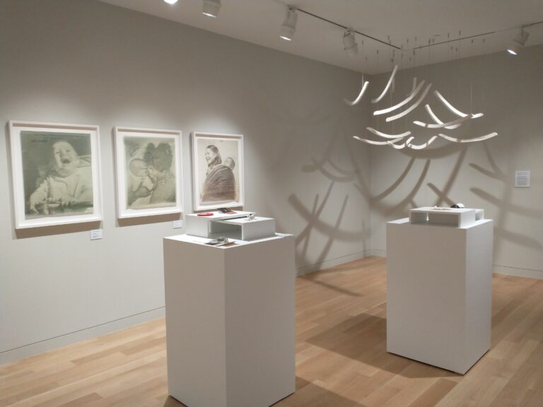 Gallery installation photo with grey walls and white pedestals with small objects on them. On the back wall are three photographs in white frames and hanging from the ceiling in the corner white curved objects.