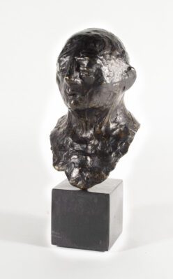 Bronze statue on a square base of a human face, bald, with closed eyes and a slightly opened mouth.
