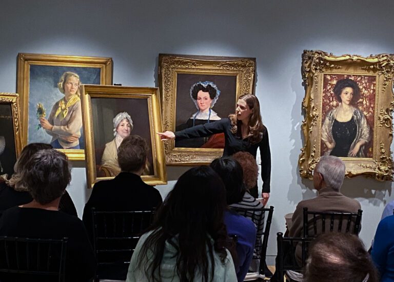 Photograph of a gallery space with many chairs and people seated for a lecture. At the front is a woman talking to the group pointing to a painting