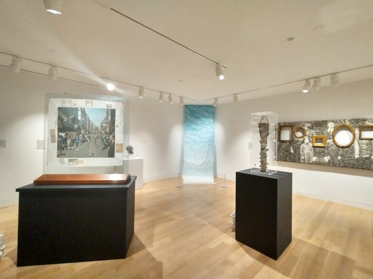 Gallery installation photo with white walls and objects on black pedestals. In the back corner is a blue fabric waterfall from ceiling to floor.