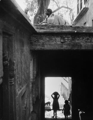 Photograph of a tunnel that leads down with a short ceiling, with two children walking down, one with a bowl on their head. On top of the tunnel is a tree and two figures sitting.