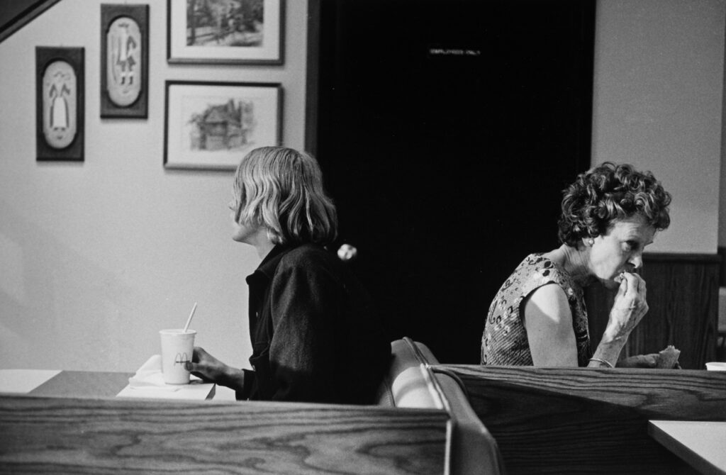 Black and white photo of two women seated back to back, eating food.