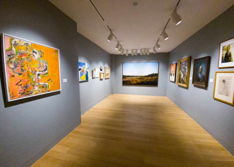 Photograph of a museum gallery room with various paintings hanging on a grey wall.