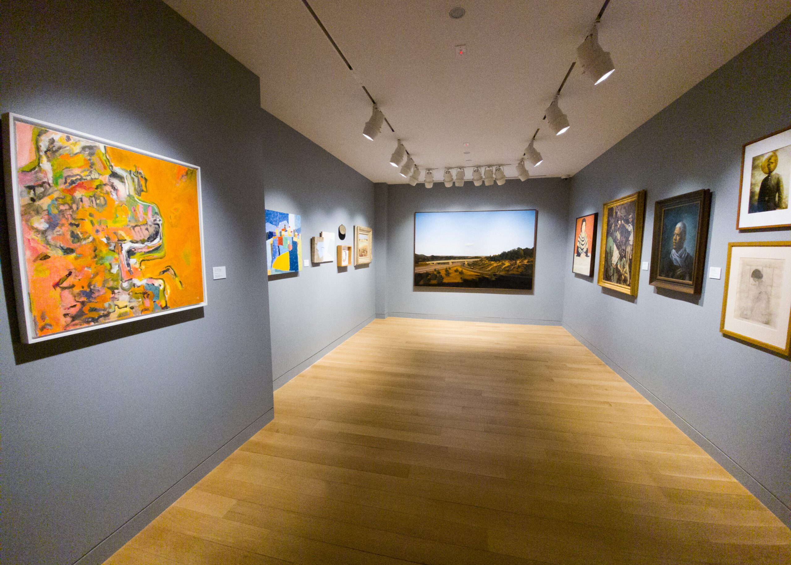 Photograph of a museum gallery room with various paintings hanging on a grey wall.