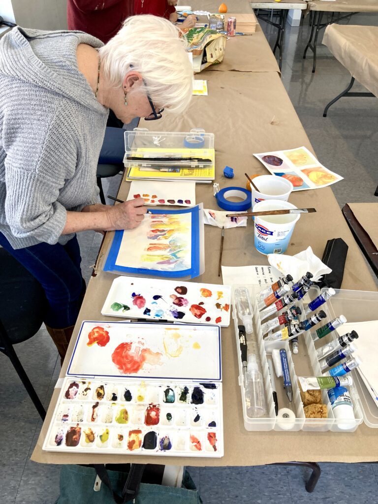 Photograph of a woman leaning over a brown paper covered table. She has short white hair, glasses, and wearing a grey hooded sweater. She has watercolor paints, palettes, and paintbrushes around her, and is painting on a white piece of paper in front of her.