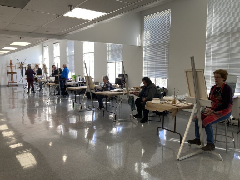 Photograph of a painting studio with seven older adults, seated or standing, along the back wall, all at easels, painting.