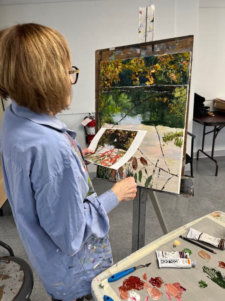 Photograph of a woman, from behind, with short brown hair wearing a blue painting smock. She is standing at an easel with a landscape painting. Her paints are to the bottom right, and she is holding her photograph inspiration near the work.