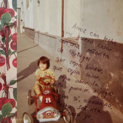 Old colored photograph of a young child seated in a red toy car driving down a side walk. Imposed over on the right is a paragraph in Portuguese, and on the far left vertically a strip of cloth with a cherry on twigs pattern.