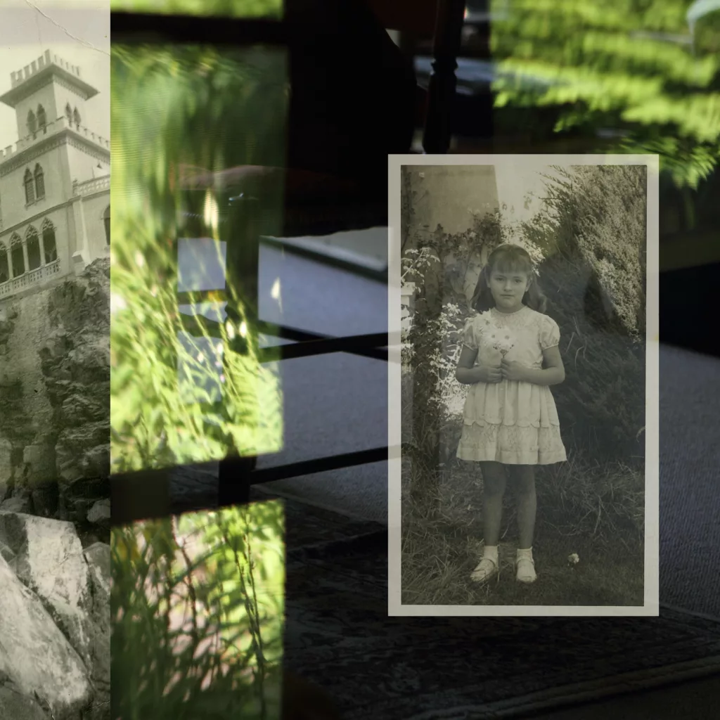 Photograph of a background containing an old building to the far left, green window reflections of grass over a shadowed interior room on the right. To the far right is a small black and white photograph of a young girl in a short dress holding a stuffed animal outside.