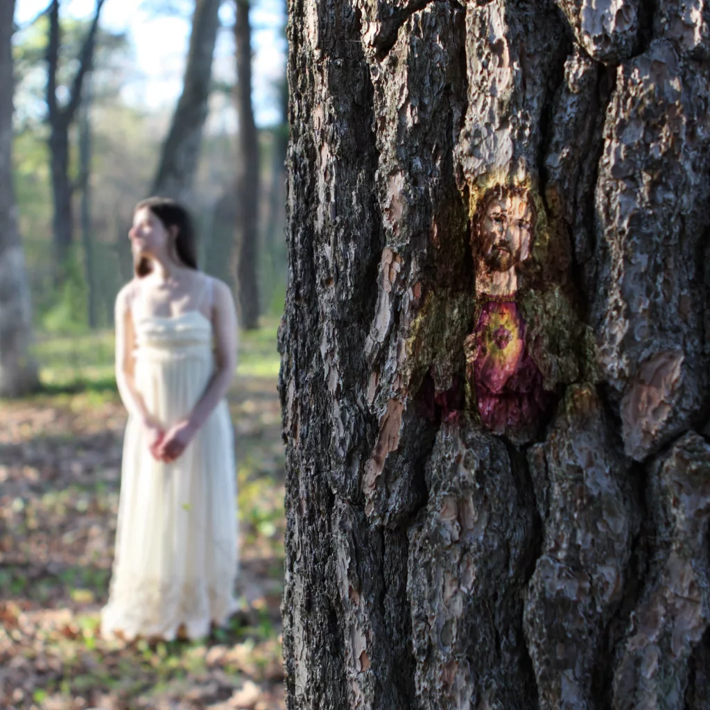 Photograph of tree bark on the right with a reflection of a painting of Jesus. Behind on the left is a wooded landscape and a light skinned woman in a plain white strap dress looking to the left and hands clasped in front of her.