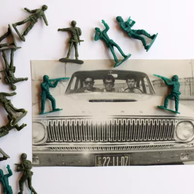 Photograph of an old back and white photograph of three men in an old car. Surrounding them are plastic green army men toys, two of which are at the passenger's head level aiming guns.