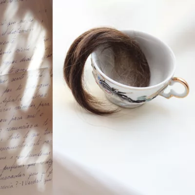 Photograph with a skinny vertical strip on the left of a hand written letter splashed with bars of light. On the right side is a white surface with a teacup holding a curling lock of hair that extends out of the teacup.