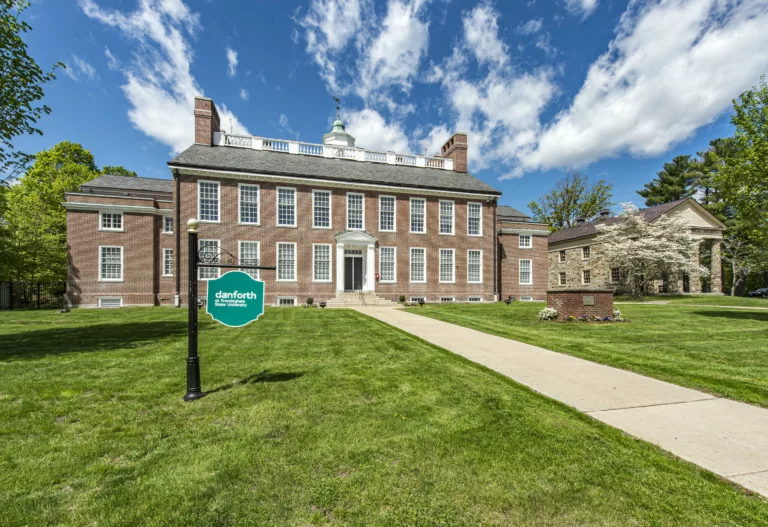 Photograph of a two story brick building at a slight angle. The windows and door are framed in white. The roof has tall brick fireplace in a rectangle on the far right, white fencing, and a cupola with a green roof in the center. There is a long green lawn with teal sign and a bright blue sky.