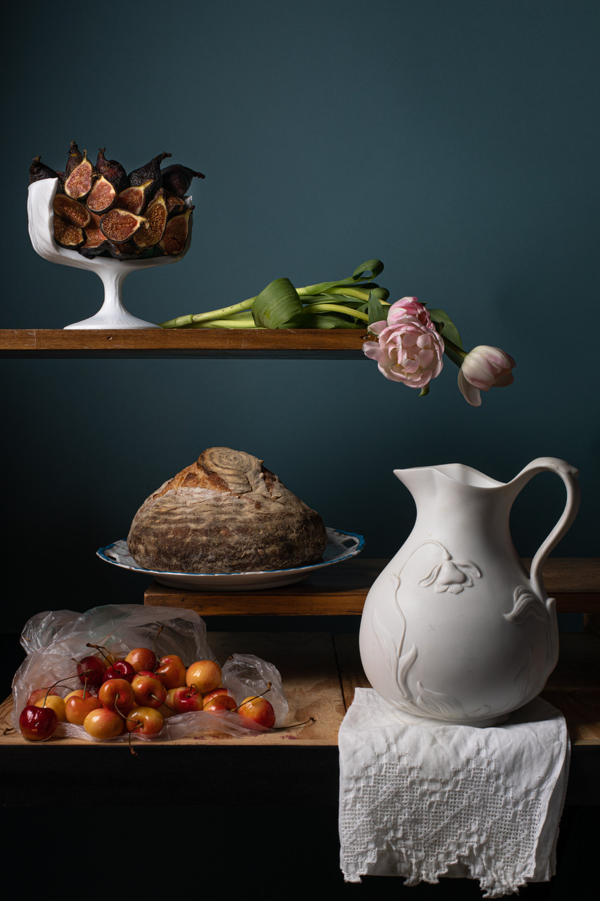Photograph of three wooden shelves in front of a deep teal-green background. The top shelf has a white broken bowl filled with figs and two pink flowers laying on their side. The middle shelf has a loaf of bread. The bottom shelf has a bag of cherries and a white ceramic pitcher.
