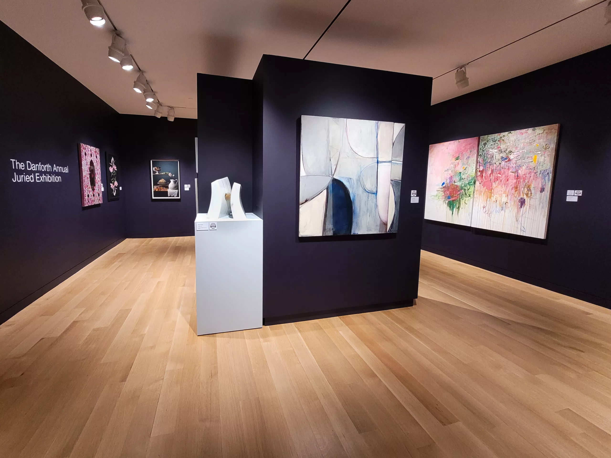 Photograph of a gallery installation with deep purple walls. There are paintings along the perimeter walls and the central wall, with one marble abstract sculpture on a light blue pedestal.