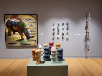 Photograph of a gallery installation with grey walls with a painting of a quilted rino on the left, and two sculptural hanging works on the center and right. In the foreground is a green pedestal with a grouping of undulating ceramics in different colors.